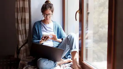 Eine Person sitzt am Fenster, hält ein Smartphone und einen Laptop. Vorhänge und ein Fensterrahmen sind sichtbar.