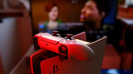 Nahaufnahme eines weißen PlayStation 5 DualSense-Controllers, der auf einer Konsole mit dem PlayStation-Logo ruht. Ein Bildschirm ist im Hintergrund zu sehen.