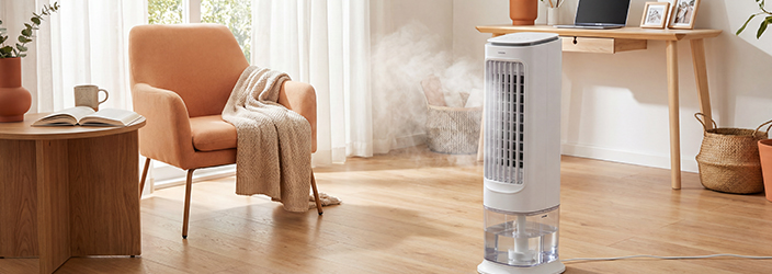 Mobiler Luftkühler mit Wassertank im modernen Wohnzimmer, sichtbar Nebel ausstrahlend. Holzschreibtisch mit Laptop, orangefarbener Lehnsessel, Naturholzmöbel und Pflanzen. Warmes, helles Ambiente mit Tageslicht.
