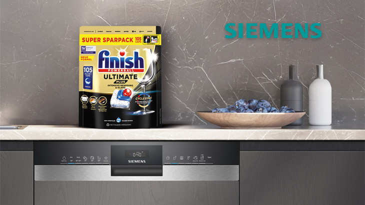 Spülmaschine unter Geschirrspülmittel, Schale mit lila Blüten, graue und weiße Flasche auf einer Marmoroberfläche.