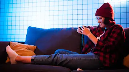 Eine Person mit roter Mütze, kariertem Hemd und Jeans sitzt auf einer Couch und benutzt ein Handy. Im Hintergrund ist eine blau beleuchtete Wand.