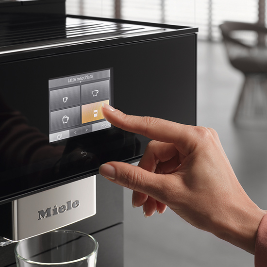 Eine Hand berührt den Bildschirm einer schwarzen Miele-Kaffeemaschine. Ein Glas steht unter dem Auslauf.