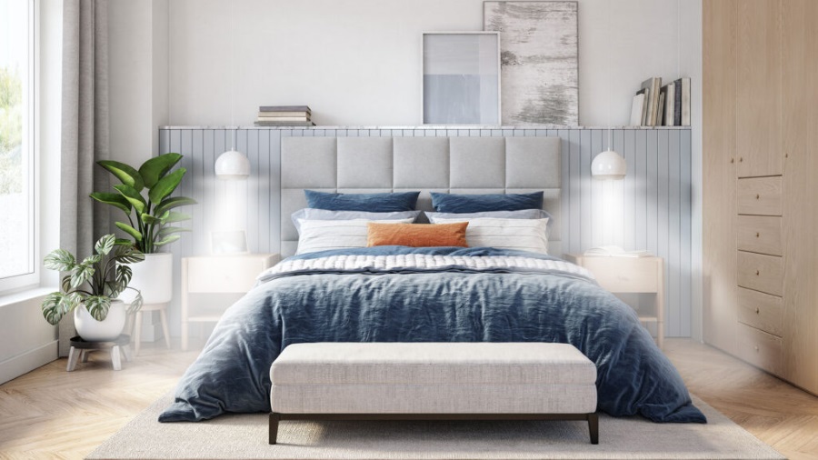 Schlafzimmer mit blau-weisser Bettwäsche, einem gepolsterten Kopfteil und Nachttischen mit Lampen. Pflanzen stehen am Fenster.