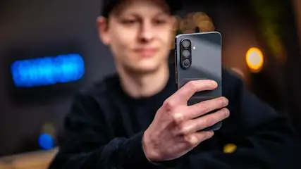 Eine Person hält ein graues Smartphone mit drei Linsen in der Hand. Im Hintergrund sind Lichter zu sehen.