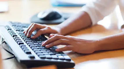 Eine Person tippt auf einer schwarzen Computertastatur auf einem hellbraunen Holztisch, mit einer Maus im Hintergrund.