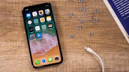 Auf einem Holztisch liegt ein Smartphone mit Ladekabel, daneben ein Buch und kleine Metallkugeln.