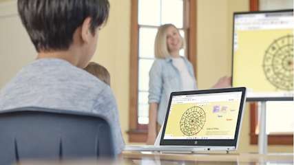 Ein Junge sitzt vor einem Laptop, im Hintergrund steht eine Lehrerin vor einer Tafel