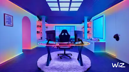 Ein Gaming-Zimmer mit Schreibtisch, Stuhl und zwei Monitoren unter blauer und rosa Beleuchtung. Ein runder Teppich liegt unter dem Tisch.