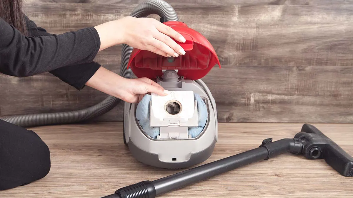 Hände einer Person entnehmen einen Staubsaugerbeutel aus einem Staubsauger mit rotem Deckel.