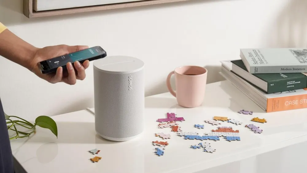 Auf einem Tisch liegen Puzzleteile, eine Tasse, Bücher und ein Lautsprecher. Eine Hand hält ein Smartphone.