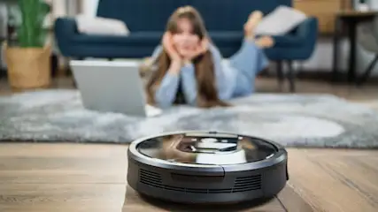 Ein Staubsaugerroboter auf Holzboden, im Hintergrund eine Frau mit Laptop auf einem Teppich vor einem blauen Sofa.