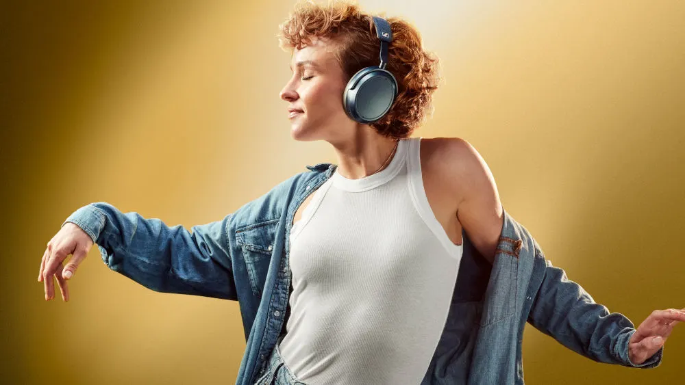 Eine rothaarige Frau in einem weißen Tanktop hört mit den Sennheiser Momentum 4 Kopfhörern Musik und tanzt dabei.