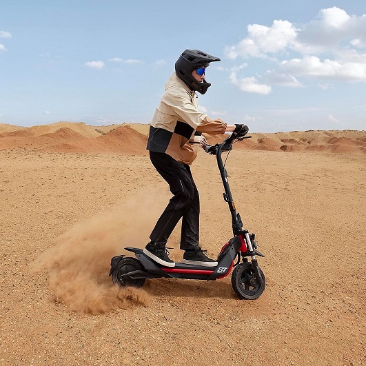 Eine Person mit Helm fährt einen roten Roller durch eine sandige Landschaft unter blauem Himmel.