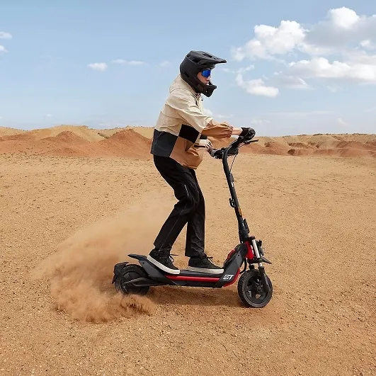 Eine Person mit Helm fährt einen roten Roller durch eine sandige Landschaft unter blauem Himmel.