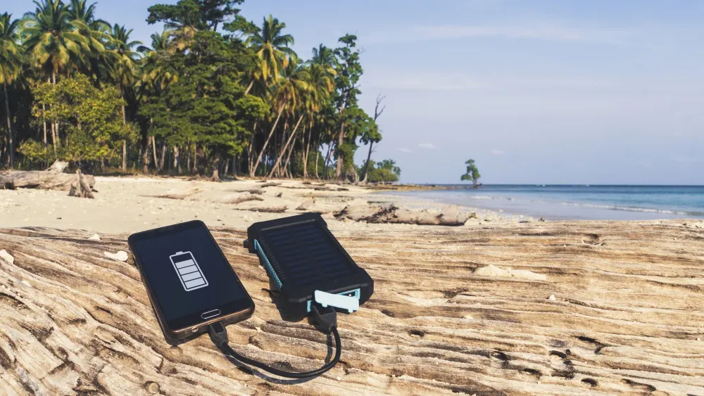 Ein Smartphone wird mit einer Powerbank am Strand mit Palmen im Hintergrund aufgeladen.