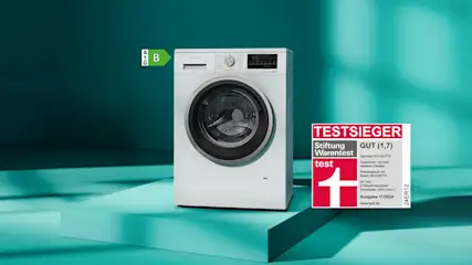 Auf blaugrünem Grund steht eine weiße Waschmaschine mit Energielabel B und ein rotes "Testsieger"-Siegel.