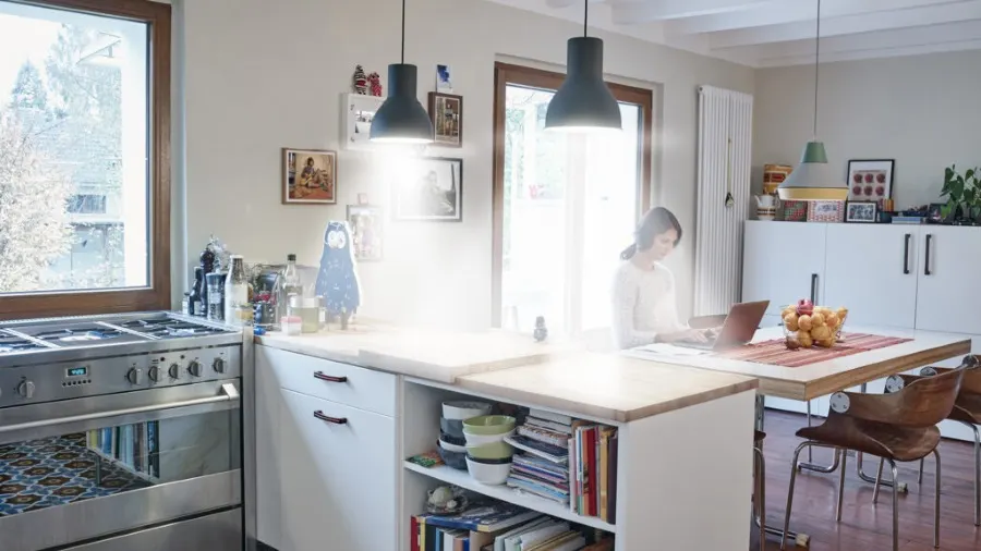 Eine Küche mit einem Herd, weißen Schränken, einer Frau am Laptop und Hängelampen. Bücher und Schüsseln sind in einem Regal.