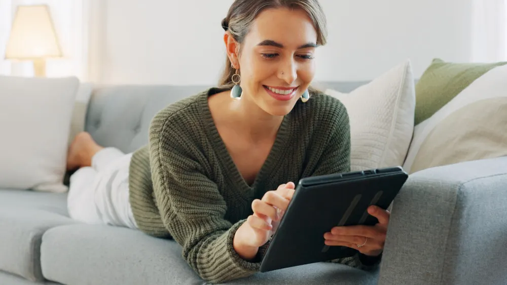 Eine Frau liegt auf einem grauen Sofa und bedient ein schwarzes Tablet. Sie trägt einen olivgrünen Pullover und Ohrringe.