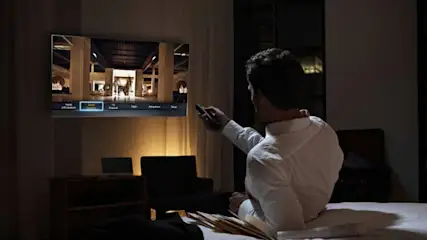 Mann mit weißem Hemd auf Bett, Fernseher an Wand zeigt Hotelfoto, Nachttischlampe leuchtet.