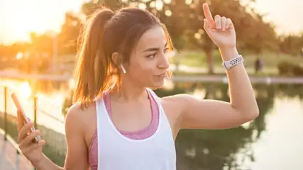 Frau mit weißem Tanktop, Arm hoch, Smartwatch, Handy in Hand. Baum und Wasser im Hintergrund.