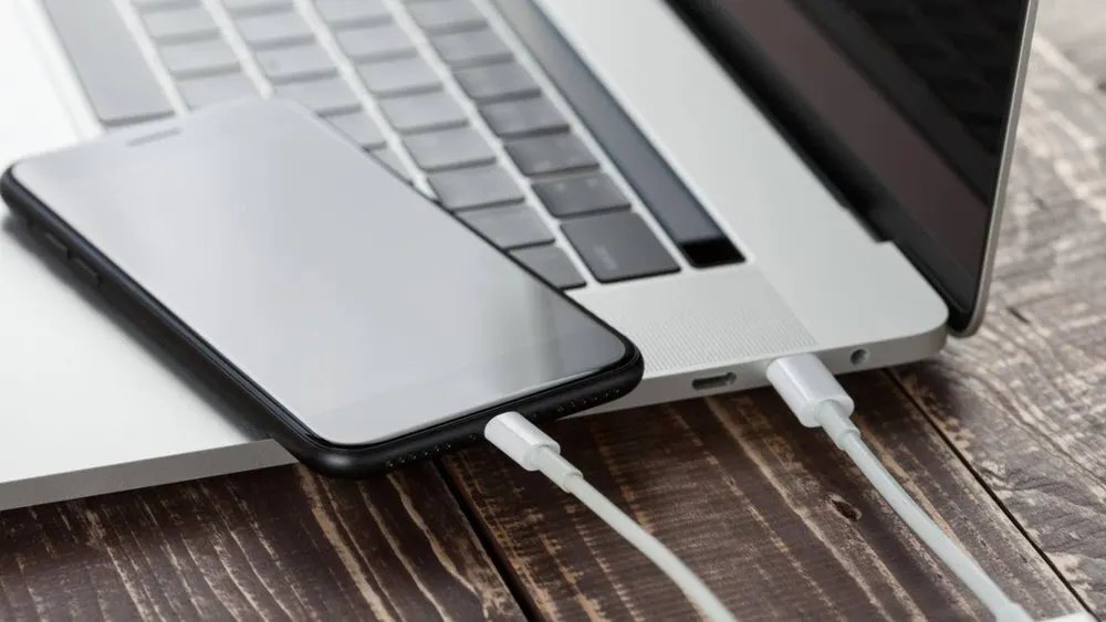 Ein Smartphone und ein Laptop liegen auf einem braunen Holztisch, verbunden durch ein weißes Ladekabel.