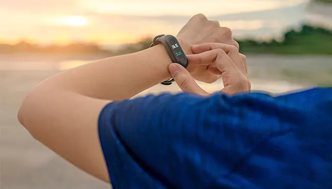 Person mit blauem Hemd, die auf eine schwarze Smartwatch mit grünem Bildschirm am Handgelenk schaut. Im Hintergrund sind Wasser und Bäume.