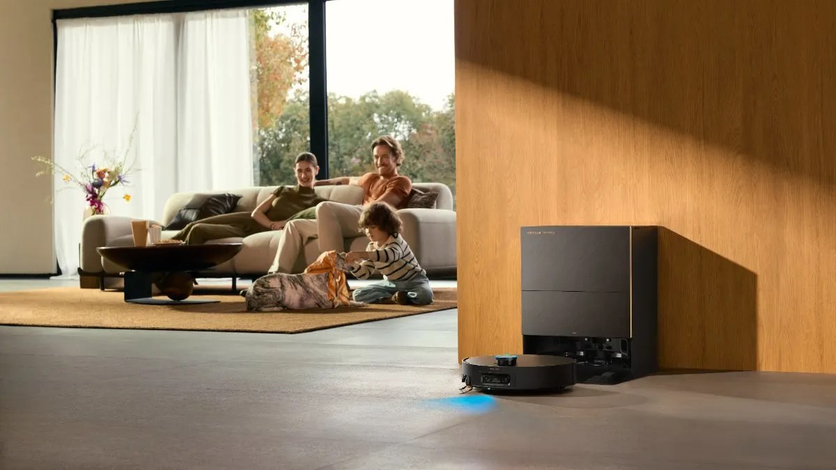 Im Hintergrund sitzen eine Frau, ein Mann und ein Junge mit Hund in einem Wohnzimmer, während sich im Vordergrund der Dreame X60 Pro Ultra Complete vor seiner Station befindet.