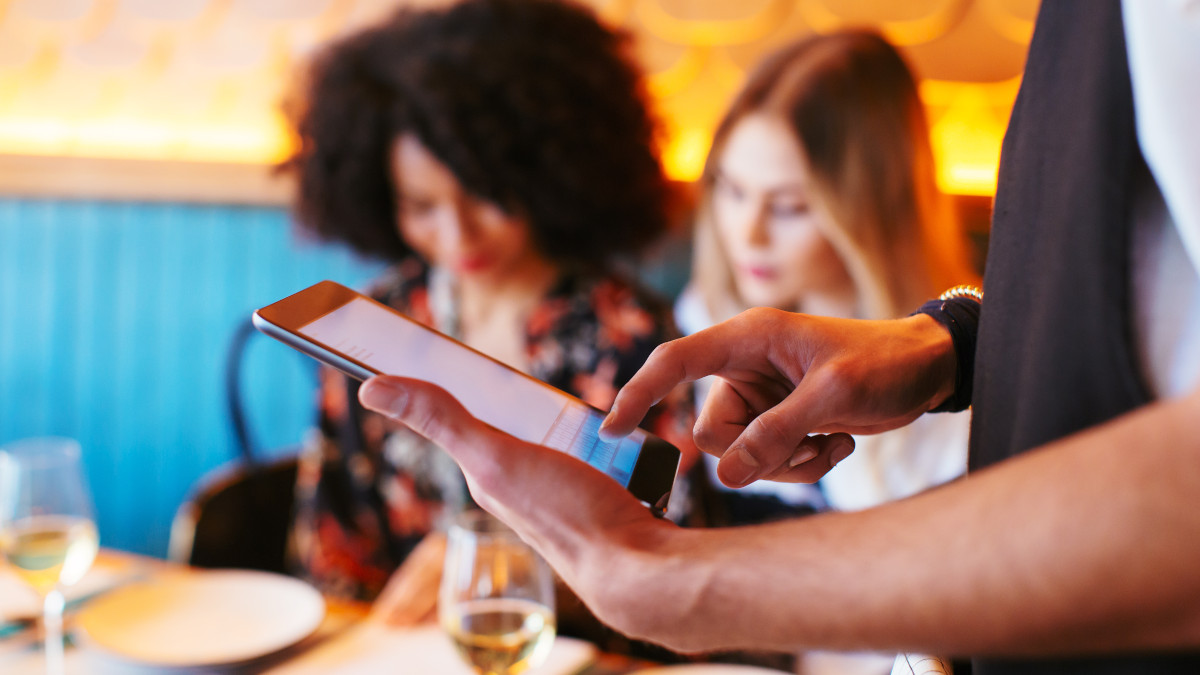 Ein Kellner bedient ein Tablet an einem Tisch mit zwei Gästen, mit Gläsern und Tellern. Der Hintergrund ist blau und orange.