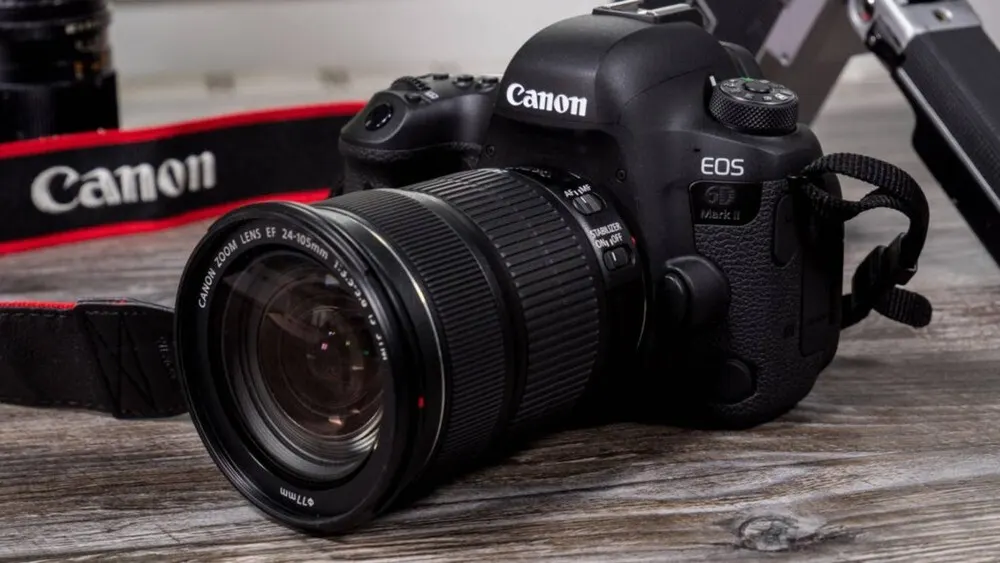 Eine schwarze Canon EOS 6D Mark II Kamera mit Objektiv liegt auf einem Holztisch, mit einem Kameragurt.