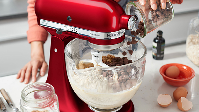 Eine rote Küchenmaschine mit Glasschüssel mischt Teig, während Schokoladenstückchen hinzugefügt werden. Eier und Zutaten sind auf der Theke.