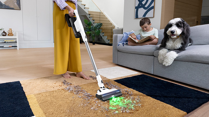 Eine Person staubsaugt einen Boden; eine Katze steht im Hintergrund auf einem Holzboden in der Nähe von Küchenschränken.