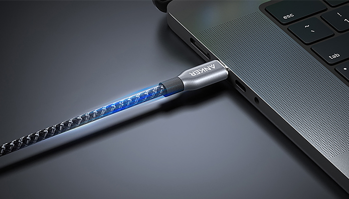 Schwarzes geflochtenes USB-Kabel mit blauer LED-Beleuchtung verbunden mit Laptop auf dunkler Oberfläche.