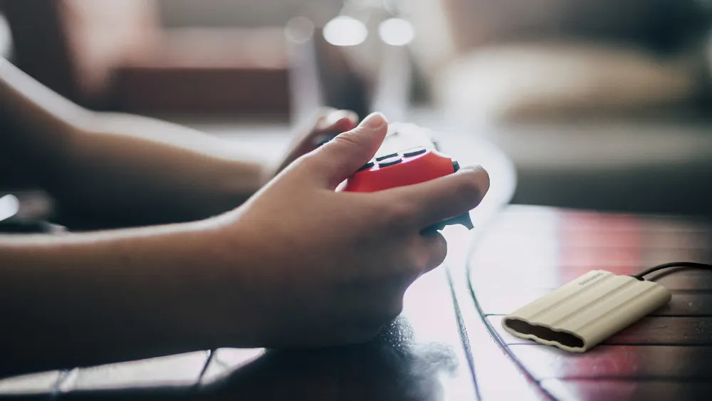 Eine Person hält einen roten Controller über einem Tisch. Eine externe Festplatte liegt daneben.