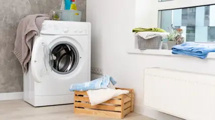 Ein Waschraum mit einer weißen Waschmaschine, einem Holzkasten mit Textilien und einem Fensterbrett mit Wäschekörben.
