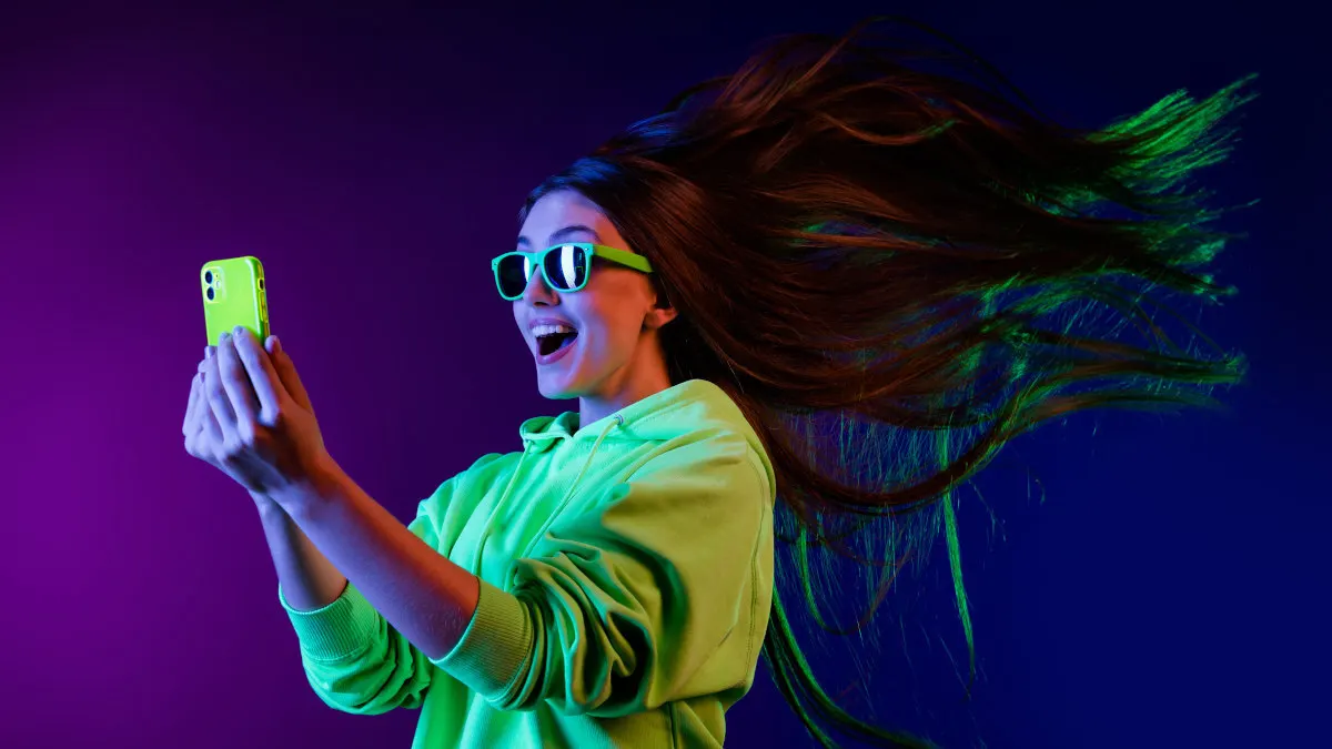 Eine Frau mit grüner Kapuzenjacke und Brille hält ein grünes Handy vor einem violetten und blauen Hintergrund.