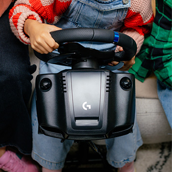 Person hält ein schwarzes Logitech-Lenkrad vor hellblauen Jeanslatzhosen und einem roten und grünen Hintergrund.