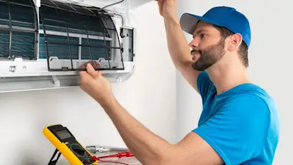 Ein Mann mit blauer Kappe repariert eine Klimaanlage. Ein gelbes Messgerät und Werkzeuge liegen darunter auf einem Tisch.