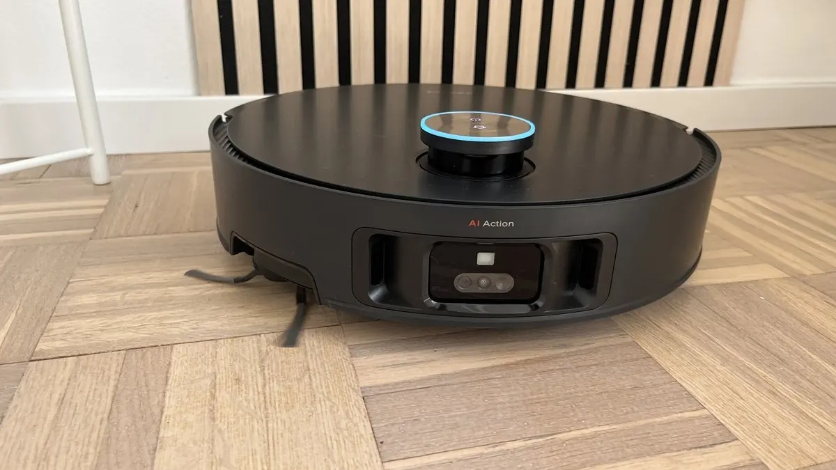 Ein runder, schwarzer Staubsaugerroboter steht auf einem hellen Holzboden vor einer Wand mit schwarzen Streifen.