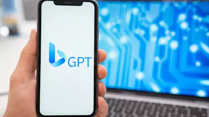 Hand hält ein Smartphone mit dem blauen "GPT"-Logo vor einem Laptop mit blauem Schaltungsdesign.