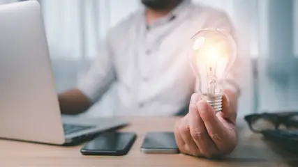 Ein Mann hält eine leuchtende Glühbirne in der Hand, daneben Laptop, Smartphones und Brille auf einem Holztisch.