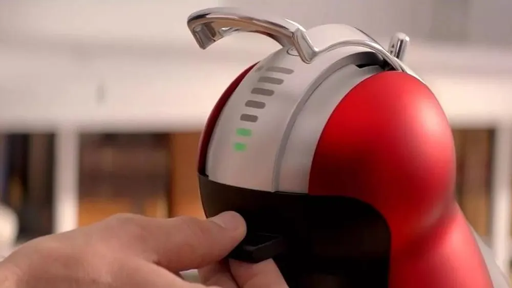 Eine Hand betätigt einen schwarzen Schalter an einer rot-weißen Kaffeemaschine mit silbernem Griff. Grüne Lichter sind sichtbar.