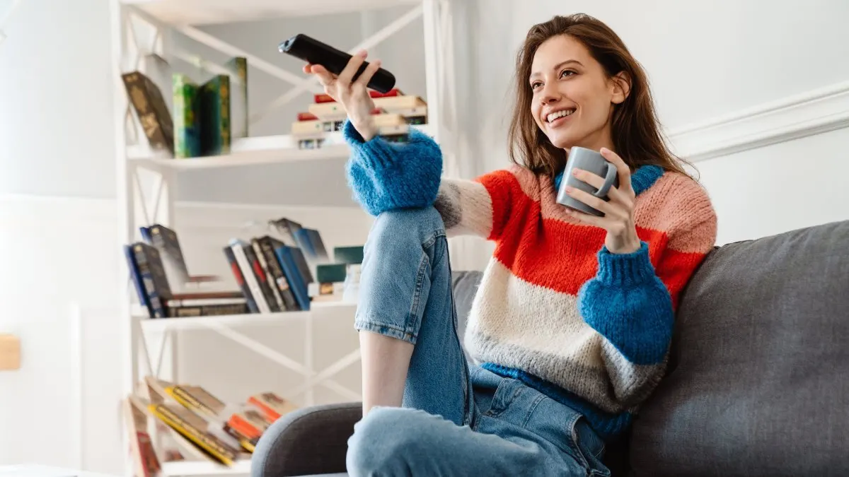 Eine Frau sitzt mit einer Fernbedienung und einer Tasse auf dem Sofa.