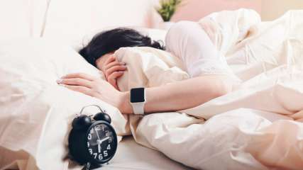 Eine Frau schläft mit einer Apple Watch am Handgelenk, neben ihr steht ein schwarzer Wecker.