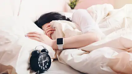 Eine Person liegt im Bett unter einer weißen Decke, das Gesicht abgewendet, mit einer schwarzen Analoguhr und einer Smartwatch.