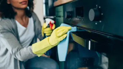 Eine Person putzt einen schwarzen Ofen mit gelben Handschuhen und einem blauen Tuch, eine Sprühflasche in der Hand.