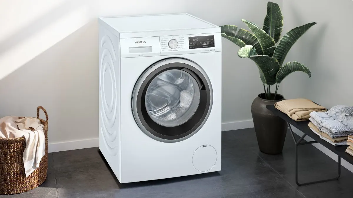 Eine weiße Siemens-Waschmaschine steht neben einem Wäschekorb und einem Tisch mit Pflanzen und Kleidung.