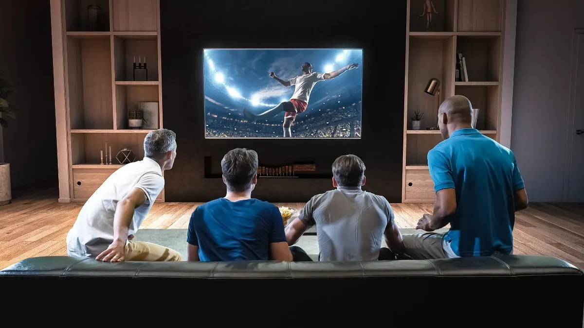 Vier Männer sitzt auf einem Sofa und schauen Fußball auf einem Fernseher.