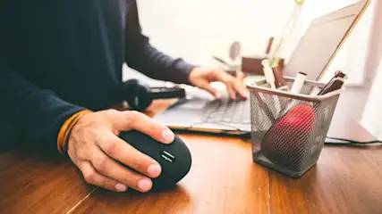Eine Person bedient eine schwarze Maus vor einem Laptop auf einem Holztisch. Ein Stiftehalter mit Stiften und ein roter Apfel sind sichtbar.