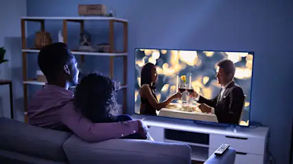 Ein Paar sitzt auf einem Sofa und schaut einen Fernseher, auf dem ein Paar beim Abendessen gezeigt wird.