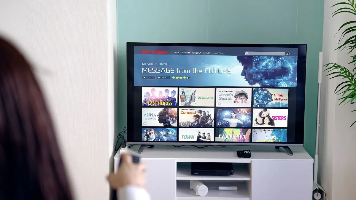 Eine Frau zeigt mit einer Fernbedienung in Richtung eines eingeschalteten Smart-TVs.